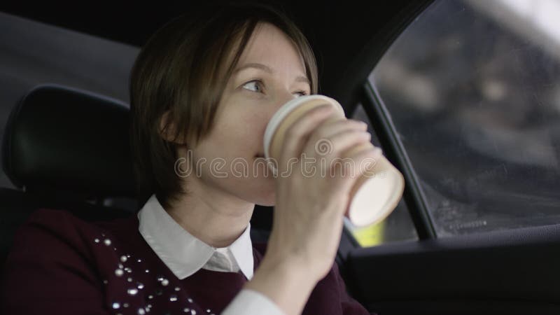 La donna graziosa con capelli marroni in blusa bianca e maglione marrone rossiccio stucking nel traffico in automobile e beve il