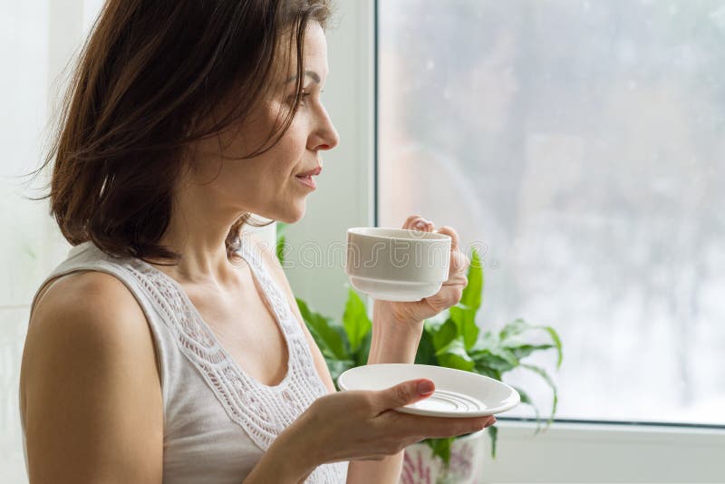La donna matura beve il caffè di mattina e guarda fuori la finestra a casa Foto in serio femminile di profilo fotografie stock La donna matura beve il caffè di mattina e guarda fuori la finestra a casa Foto in serio femminile di profilo fotografie stock