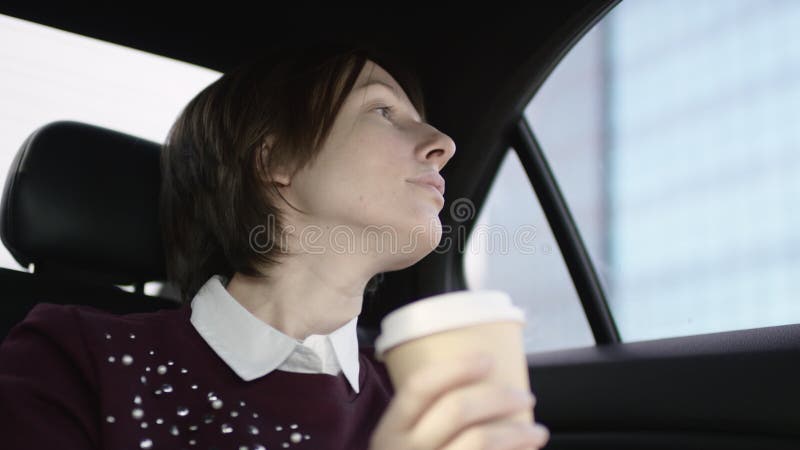 La donna piacevole con capelli marroni in blusa bianca e maglione marrone rossiccio stucking nel traffico in automobile e beve il