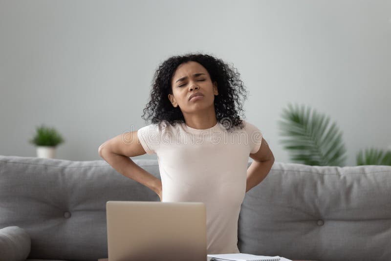 Mixed-race unhealthy african woman sitting on sofa touches lower back suffers from severe pain caused by overuse of computer lot of time working on pc, poor posture and improper spinal support concept. Mixed-race unhealthy african woman sitting on sofa touches lower back suffers from severe pain caused by overuse of computer lot of time working on pc, poor posture and improper spinal support concept