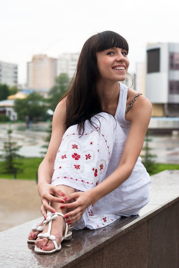La donna sorridente attraente si siede sopra il fondo della città fotografie stock La donna sorridente attraente si siede sopra il fondo della città fotografie stock