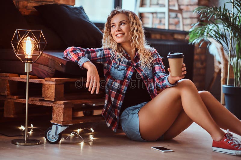 La donna sorridente graziosa sta sedendosi accanto al sof? del pallet Sta tenendo la tazza di caff? immagine stock La donna sorridente graziosa sta sedendosi accanto al sof? del pallet Sta tenendo la tazza di caff? immagine stock