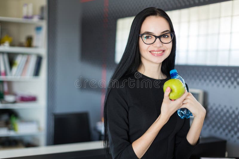 La empresaria joven hermosa en vestido negro y los vidrios se sientan en la tabla en la oficina y sostienen la manzana y la botel imágenes de archivo libres de regalías La empresaria joven hermosa en vestido negro y los vidrios se sientan en la tabla en la oficina y sostienen la manzana y la botel imágenes de archivo libres de regalías