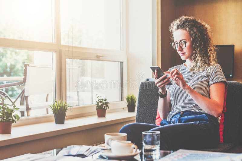 La empresaria joven se sienta en el sofá cerca de ventana, utiliza smartphone Muchacha que charla, blogging, comprobando el corre fotografía de archivo libre de regalías La empresaria joven se sienta en el sofá cerca de ventana, utiliza smartphone Muchacha que charla, blogging, comprobando el corre fotografía de archivo libre de regalías