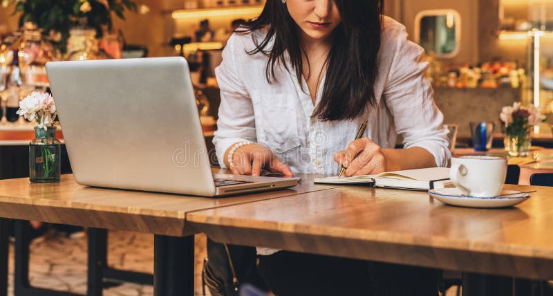 La empresaria se está sentando en café en la tabla delante del ordenador portátil, haciendo notas en el cuaderno, trabajando Estu imagen de archivo La empresaria se está sentando en café en la tabla delante del ordenador portátil, haciendo notas en el cuaderno, trabajando Estu imagen de archivo