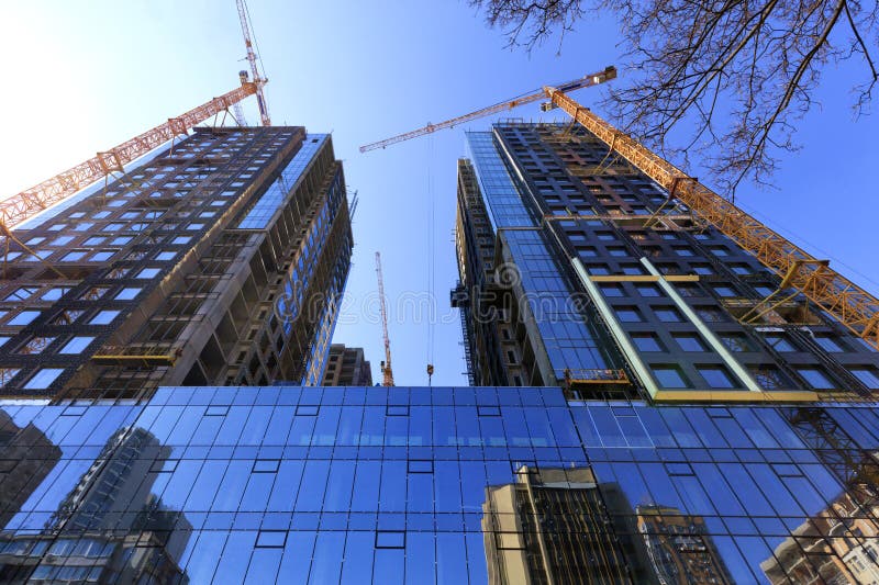 La facciata di vetro, una riflessione del cielo blu e gru vicino ad una costruzione concreta moderna in costruzione fotografia stock La facciata di vetro, una riflessione del cielo blu e gru vicino ad una costruzione concreta moderna in costruzione fotografia stock