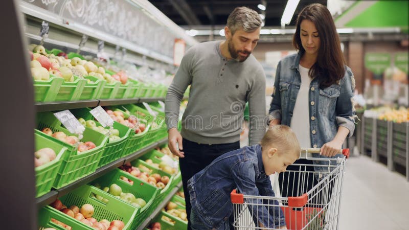 La familia joven con el niño está haciendo compras para la comida en supermercado, los padres están eligiendo la fruta y el mucha