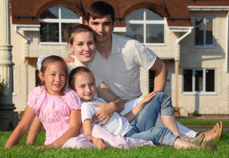 La familia a partir del cuatro se sienta en hierba contra casa imagen de archivo libre de regalías La familia a partir del cuatro se sienta en hierba contra casa imagen de archivo libre de regalías