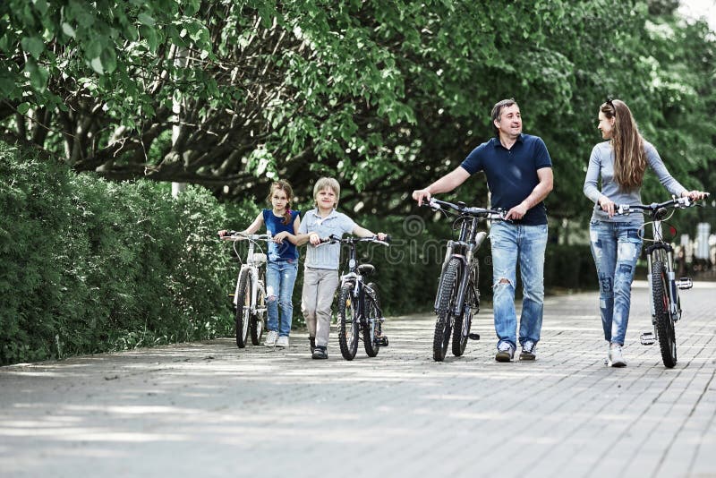 La famille moderne avec leurs vélos marchent par le parc de ville photographie stock La famille moderne avec leurs vélos marchent par le parc de ville photographie stock
