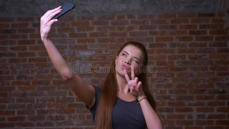 La femelle caucasienne mignonne prend le selfie et montre ses deux doigts à son téléphone tout en se tenant sur le fond de brique