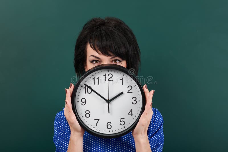 La femme cache son visage derrière l'horloge, posant par le panneau de craie, le temps et le concept d'éducation, fond vert, tir photos stock La femme cache son visage derrière l'horloge, posant par le panneau de craie, le temps et le concept d'éducation, fond vert, tir photos stock