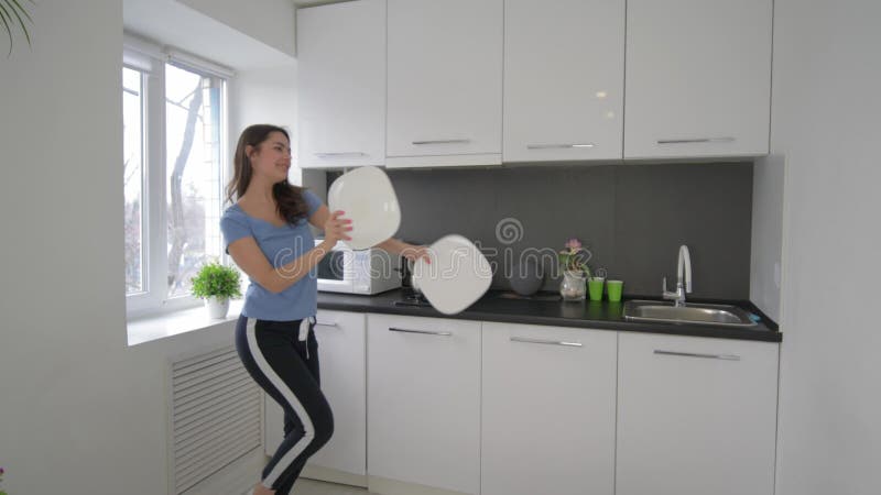 La femme drôle heureuse de femme au foyer dupant autour et chante avec des plats dans des bras à la cuisine à la maison