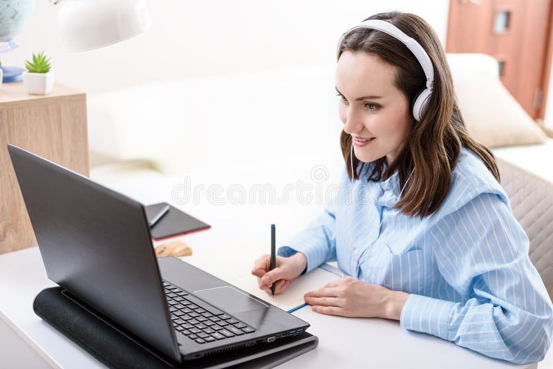 La femme du caucase avec des écouteurs écrit dans les regards du carnet au travail à distance d'écran d'ordinateur portable à la m photo stock
