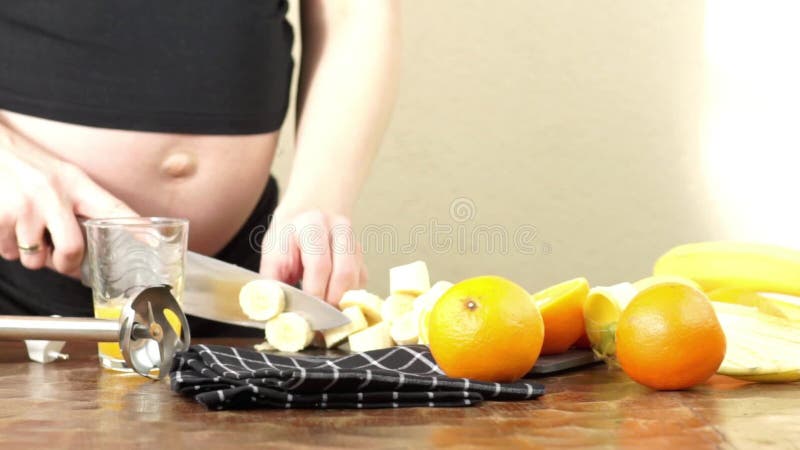 La femme enceinte fait un smoothie sain dans ses tranches de cuisine viennent des bananes et des oranges banque de vidéos