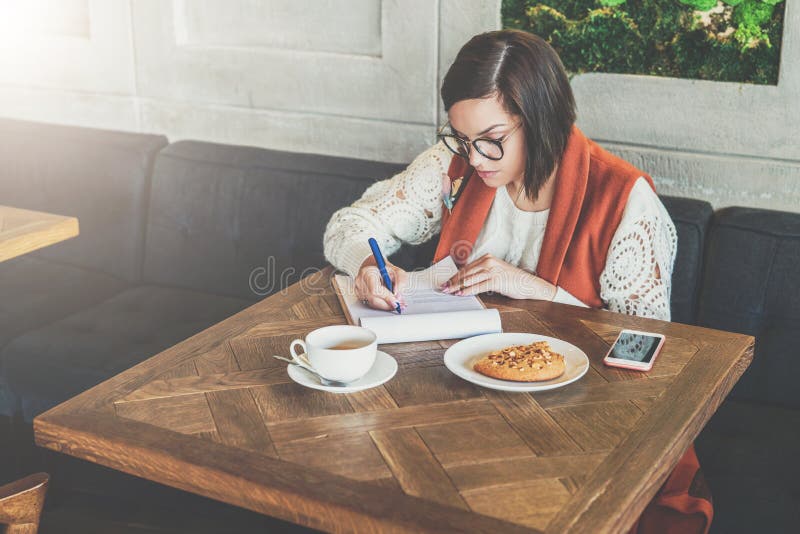La femme s'assied en café à la table La fille complète une application, questionnaire, documents de signes, élabore le résumé photographie stock La femme s'assied en café à la table La fille complète une application, questionnaire, documents de signes, élabore le résumé photographie stock