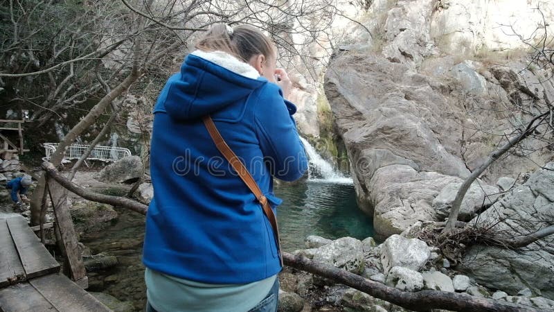 La femme se tient sur le pont en bois au-dessus de la rivière dans la forêt, prenant des photos clips vidéos