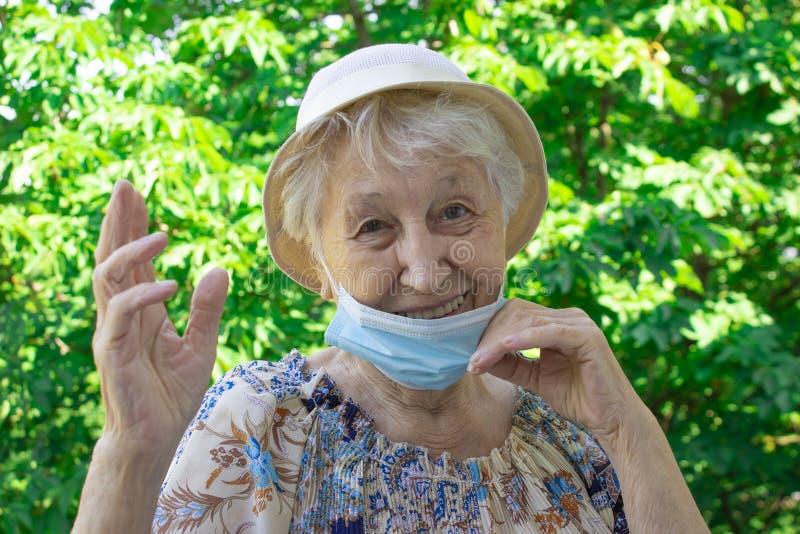 Positive smiling senior woman takes off protective medical mask from face at balcony. Concept of happy end or victory over coronavirus. Pandemic Covid-19. Green trees at summer. Positive smiling senior woman takes off protective medical mask from face at balcony. Concept of happy end or victory over coronavirus. Pandemic Covid-19. Green trees at summer