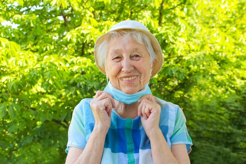 Positive smiling senior woman takes off protective medical mask from face outdoors. Concept of happy end. Pollen allergy. Victory over coronavirus. Pandemic Covid-19. Green trees at summer. Positive smiling senior woman takes off protective medical mask from face outdoors. Concept of happy end. Pollen allergy. Victory over coronavirus. Pandemic Covid-19. Green trees at summer