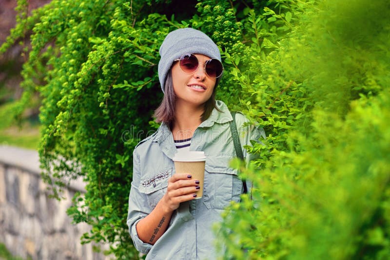 La femmina in occhiali da sole tiene la tazza di caffè di carta in un parco dell'estate
