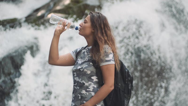 La fille attirante boit l'eau froide d'une bouteille et éteint la soif tout en se tenant sur une pierre près d'une cascade C? t? banque de vidéos