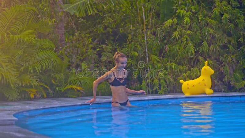 La fille blonde entre dans l'eau à partir du bord de piscine à l'encontre les plantes vertes