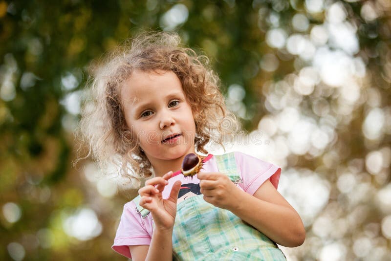 La fille blonde mignonne joue avec le chesnut en parc, feuilles d'automne colorées à l'arrière-plan, enfant gai, extérieur, heure images libres de droits La fille blonde mignonne joue avec le chesnut en parc, feuilles d'automne colorées à l'arrière-plan, enfant gai, extérieur, heure images libres de droits