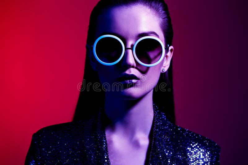 La fille de mode avec de longs cheveux et lunettes de soleil rondes dans une robe brillante noire pose dans la lampe au néon dans photographie stock La fille de mode avec de longs cheveux et lunettes de soleil rondes dans une robe brillante noire pose dans la lampe au néon dans photographie stock
