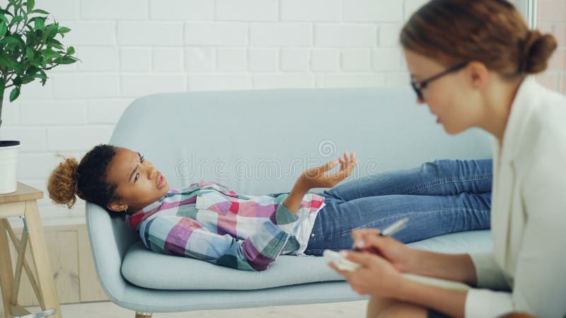 La fille déprimée d'Afro-américain parle au psychologue se trouvant sur le sofa et parlant tandis que le thérapeute en verres est clips vidéos