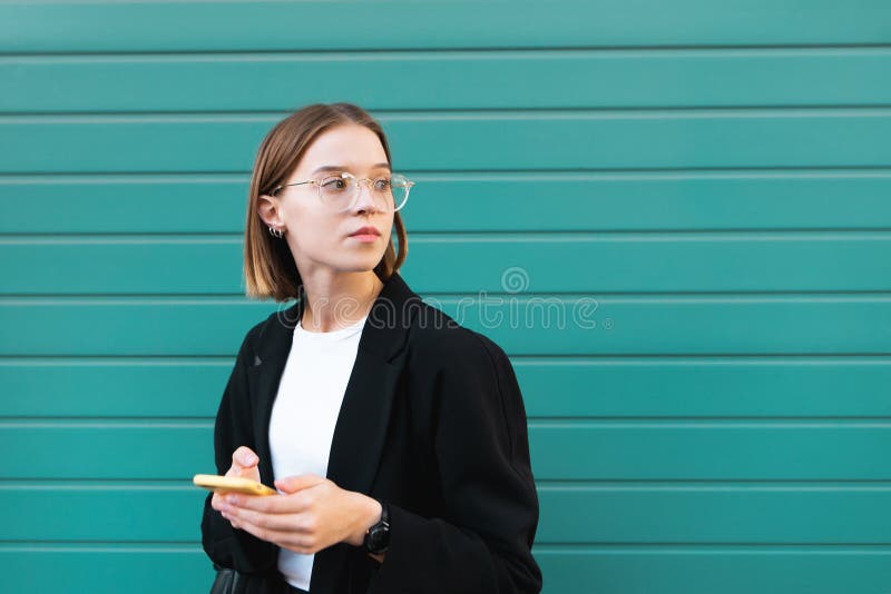 La fille en veste et verres, se tient avec un smartphone dans des ses mains sur un fond de turquoise photos libres de droits La fille en veste et verres, se tient avec un smartphone dans des ses mains sur un fond de turquoise photos libres de droits