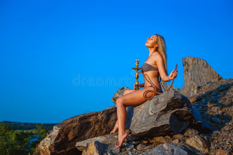 Girl dressed in a bikini smokes a hookah on a hillside. Relax and blue sky. Girl dressed in a bikini smokes a hookah on a hillside. Relax and blue sky.