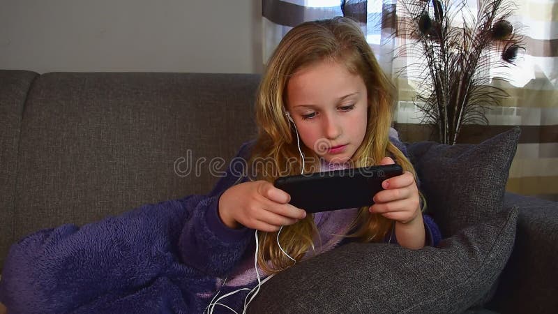 La fille mignonne de 9 ans se trouve sur le divan de ses jeux de pyjama avec le téléphone banque de vidéos