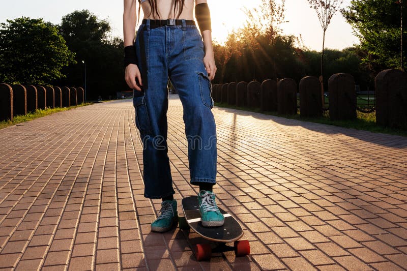 La fille mince avec des dreadlocks en jeans et monte sur un longboard au coucher du soleil photographie stock libre de droits
