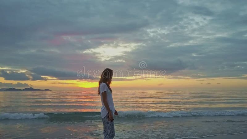 La fille mince grande va sur le ressac de plage à l'encontre le lever de soleil d'océan