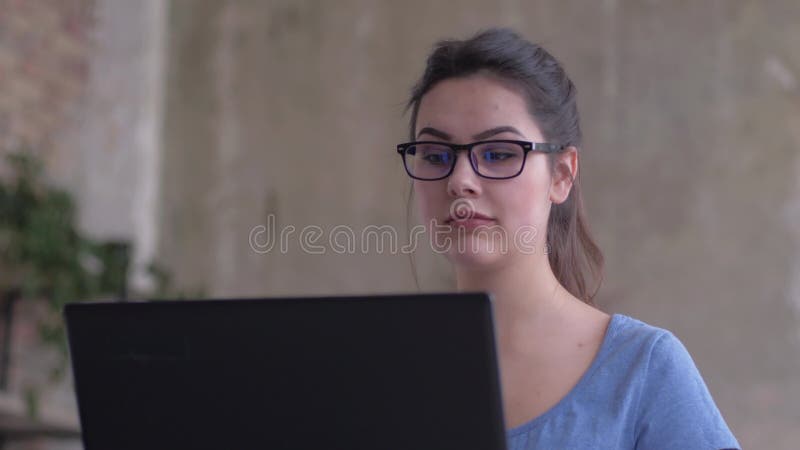 La fille moderne dans des lunettes regarde dans l'écran d'ordinateur portable et les types textotent sur le clavier pendant le p