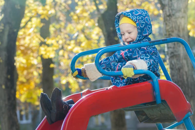 La fille riante dans la veste à capuchon chaude glissant vers le bas le terrain de jeu en plastique rouge glissent au fond jaune photo stock