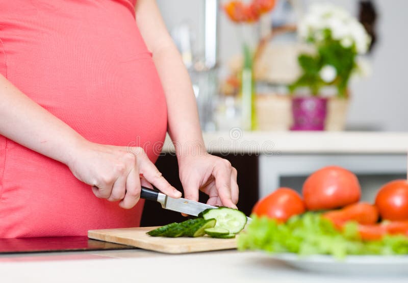 La fine sulla donna incinta con il coltello sulla cucina taglia il cetriolo fotografia stock La fine sulla donna incinta con il coltello sulla cucina taglia il cetriolo fotografia stock