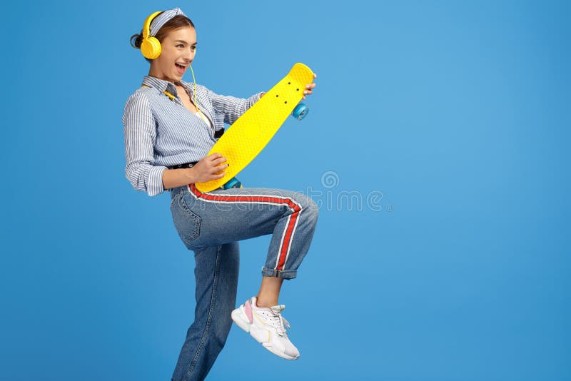 La foto de la mujer joven alegre con los auriculares amarillos juega la guitarra imaginaria con el penique o el monopatín sobre f imagen de archivo libre de regalías La foto de la mujer joven alegre con los auriculares amarillos juega la guitarra imaginaria con el penique o el monopatín sobre f imagen de archivo libre de regalías