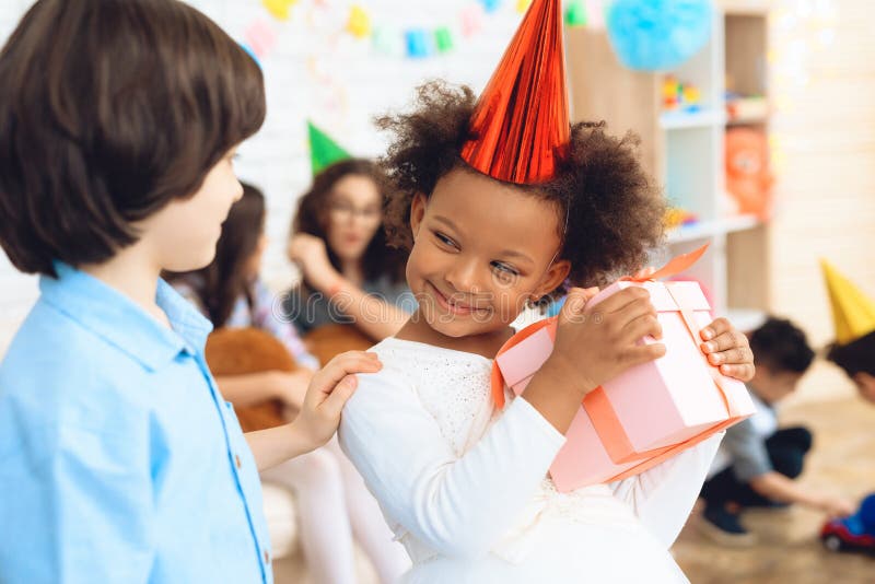 La gentille petite fille dans la robe blanche est satisfaite avec le cadeau qu'elle a été donnée par le garçon dans la chemise bl image libre de droits La gentille petite fille dans la robe blanche est satisfaite avec le cadeau qu'elle a été donnée par le garçon dans la chemise bl image libre de droits