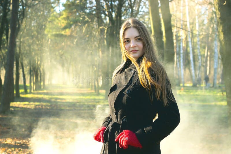 La giovane bella ragazza nei guanti rossi di un cappotto nero che esplora il fumo della foresta della molla parcheggia fotografie stock La giovane bella ragazza nei guanti rossi di un cappotto nero che esplora il fumo della foresta della molla parcheggia fotografie stock