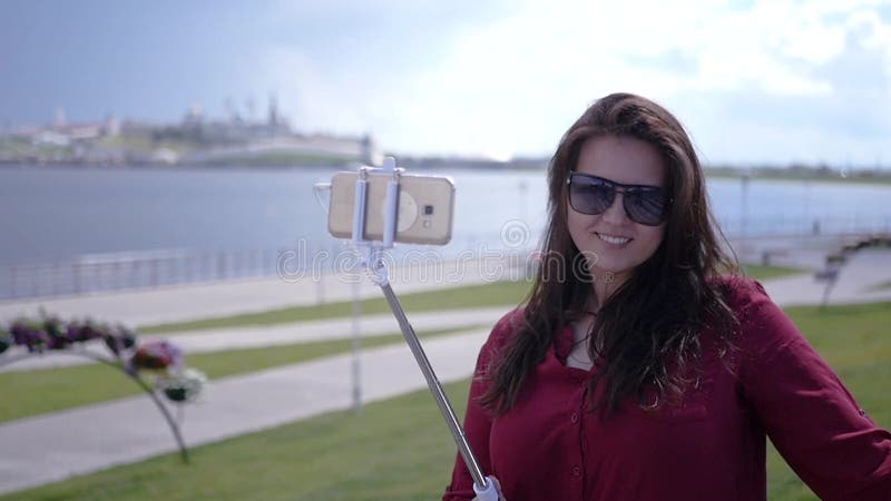 La giovane bella ragazza sta facendo una foto su un bastone del selfie su un fondo dell'acqua, della città e del cielo HD, 1920x1 stock footage La giovane bella ragazza sta facendo una foto su un bastone del selfie su un fondo dell'acqua, della città e del cielo HD, 1920x1 stock footage
