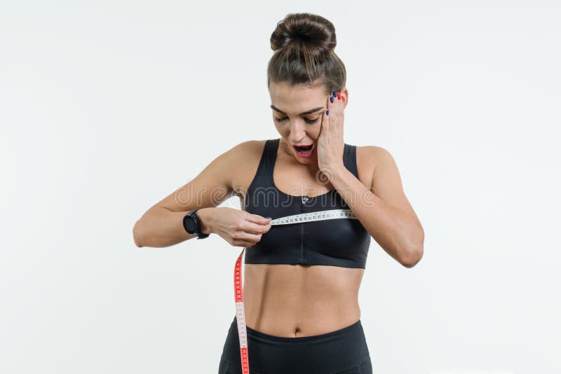 La giovane donna in abiti sportivi è sorpresa che tiene un nastro di centimetro sul suo petto il concetto dello sport, della form fotografia stock libera da diritti La giovane donna in abiti sportivi è sorpresa che tiene un nastro di centimetro sul suo petto il concetto dello sport, della form fotografia stock libera da diritti