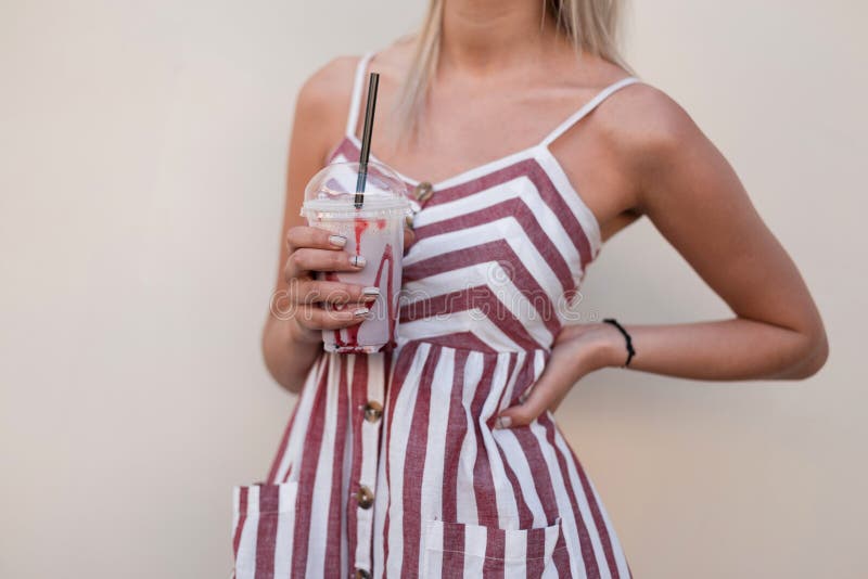 La giovane donna alla moda abbronzata in un vestito rosa a strisce alla moda posa vicino ad una parete nella città e nelle tenute immagine stock libera da diritti La giovane donna alla moda abbronzata in un vestito rosa a strisce alla moda posa vicino ad una parete nella città e nelle tenute immagine stock libera da diritti