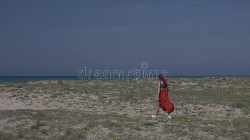 La giovane donna dai capelli rossi del movimento lento in una gonna rossa cammina lungo la riva verso il mare contro un cielo blu