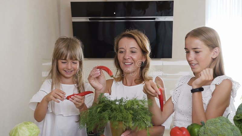 La giovane donna e due ragazze vestite in vestiti bianchi stanno giocando con peperoncino ed aneto verde a casa nella cucina archivi video