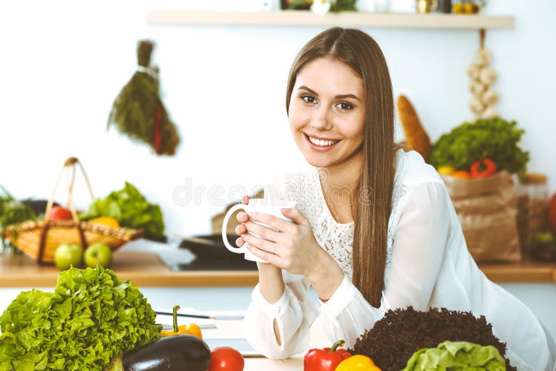 La giovane donna felice sta tenendo la tazza bianca e sta esaminando la macchina fotografica mentre si sedeva alla tavola di legn fotografia stock libera da diritti La giovane donna felice sta tenendo la tazza bianca e sta esaminando la macchina fotografica mentre si sedeva alla tavola di legn fotografia stock libera da diritti