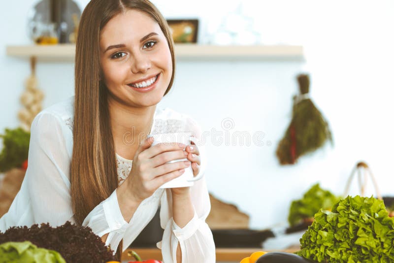 La giovane donna felice sta tenendo la tazza bianca e sta esaminando la macchina fotografica mentre si sedeva alla tavola di legn immagini stock La giovane donna felice sta tenendo la tazza bianca e sta esaminando la macchina fotografica mentre si sedeva alla tavola di legn immagini stock
