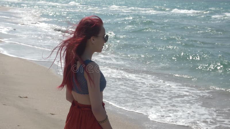 La giovane donna redheaded del movimento lento in una gonna rossa sta stando sulla spiaggia e sta esaminando la spuma