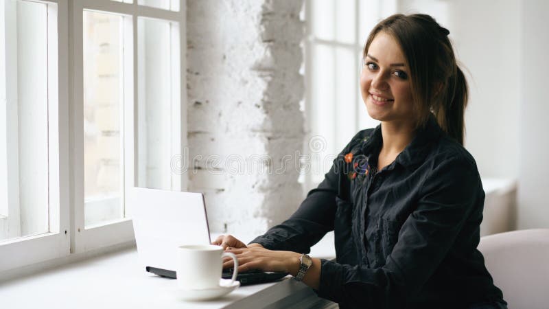La giovane donna sorridente dello studente si siede in caffetteria alla tavola con il computer portatile all'interno immagine stock libera da diritti La giovane donna sorridente dello studente si siede in caffetteria alla tavola con il computer portatile all'interno immagine stock libera da diritti