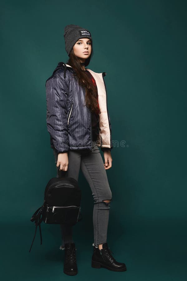 La giovane ragazza teenager castana, in rivestimento e cappello con una borsa, sta integrale sopra fondo verde scuro, immagini stock La giovane ragazza teenager castana, in rivestimento e cappello con una borsa, sta integrale sopra fondo verde scuro, immagini stock