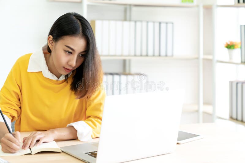 La giovane studentessa asiatica attraente che si siede alla tavola che esamina il giornale di scrittura del computer portatile no immagini stock La giovane studentessa asiatica attraente che si siede alla tavola che esamina il giornale di scrittura del computer portatile no immagini stock